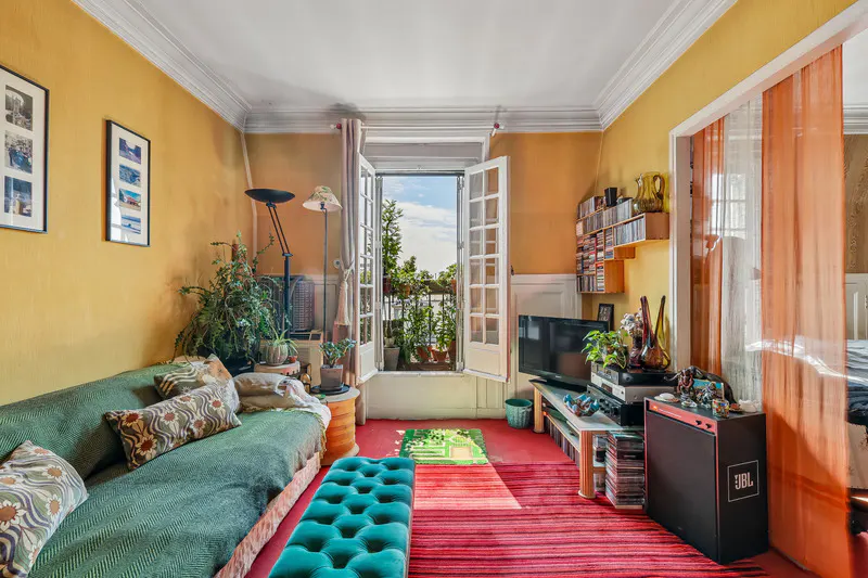 Cozy living room with yellow walls, a green sofa, and a turquoise ottoman. Open balcony doors reveal plants and a view. Red rug and eclectic decor.