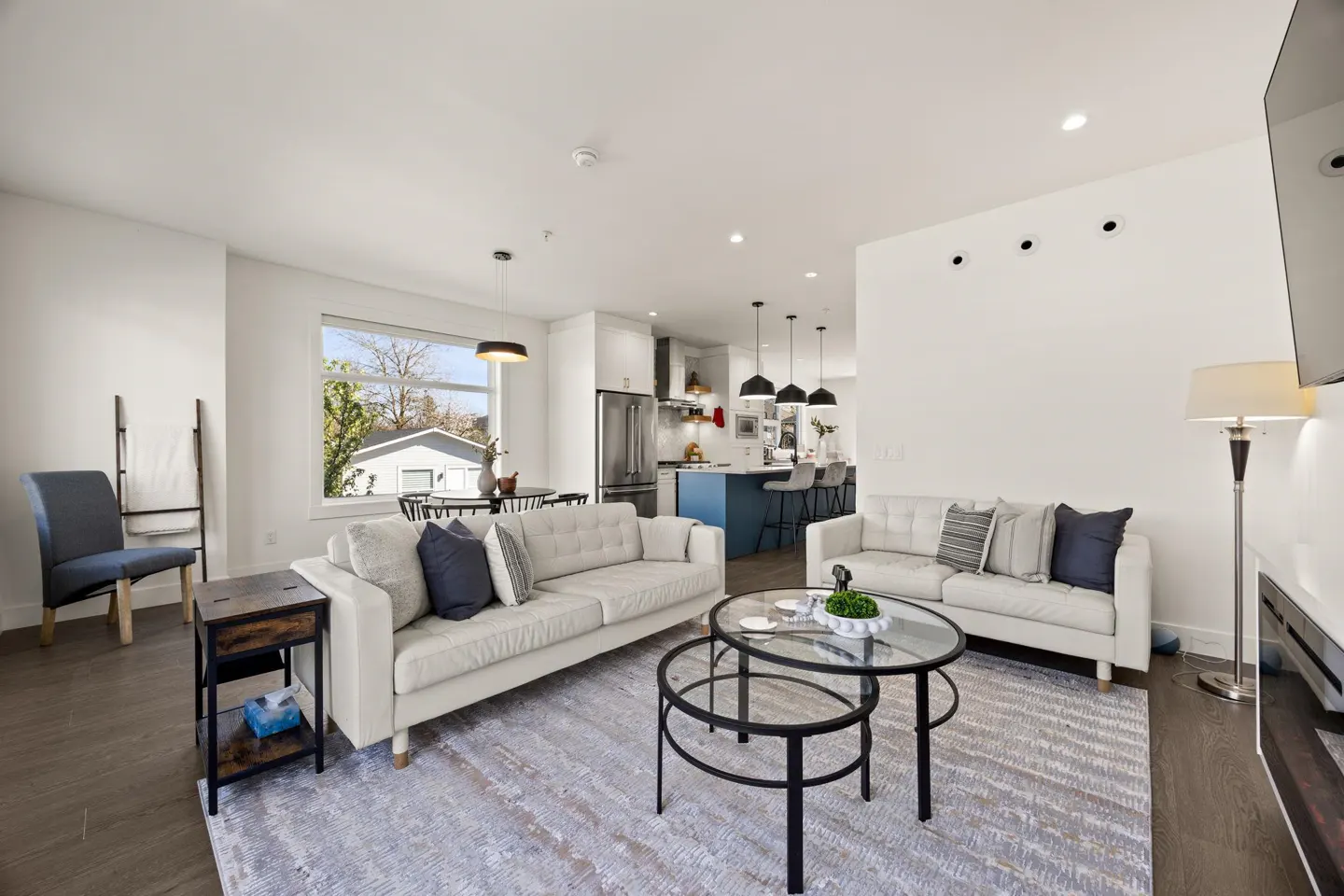 Bright, open-concept living room with white sofas, round glass tables, and a view into the kitchen with a blue island.