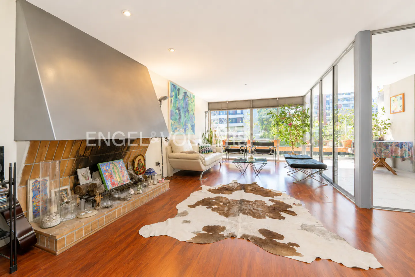 A bright living room with a brick fireplace, a white sofa, and a cowhide rug on a wood floor. A glass wall leads to a balcony with plants.