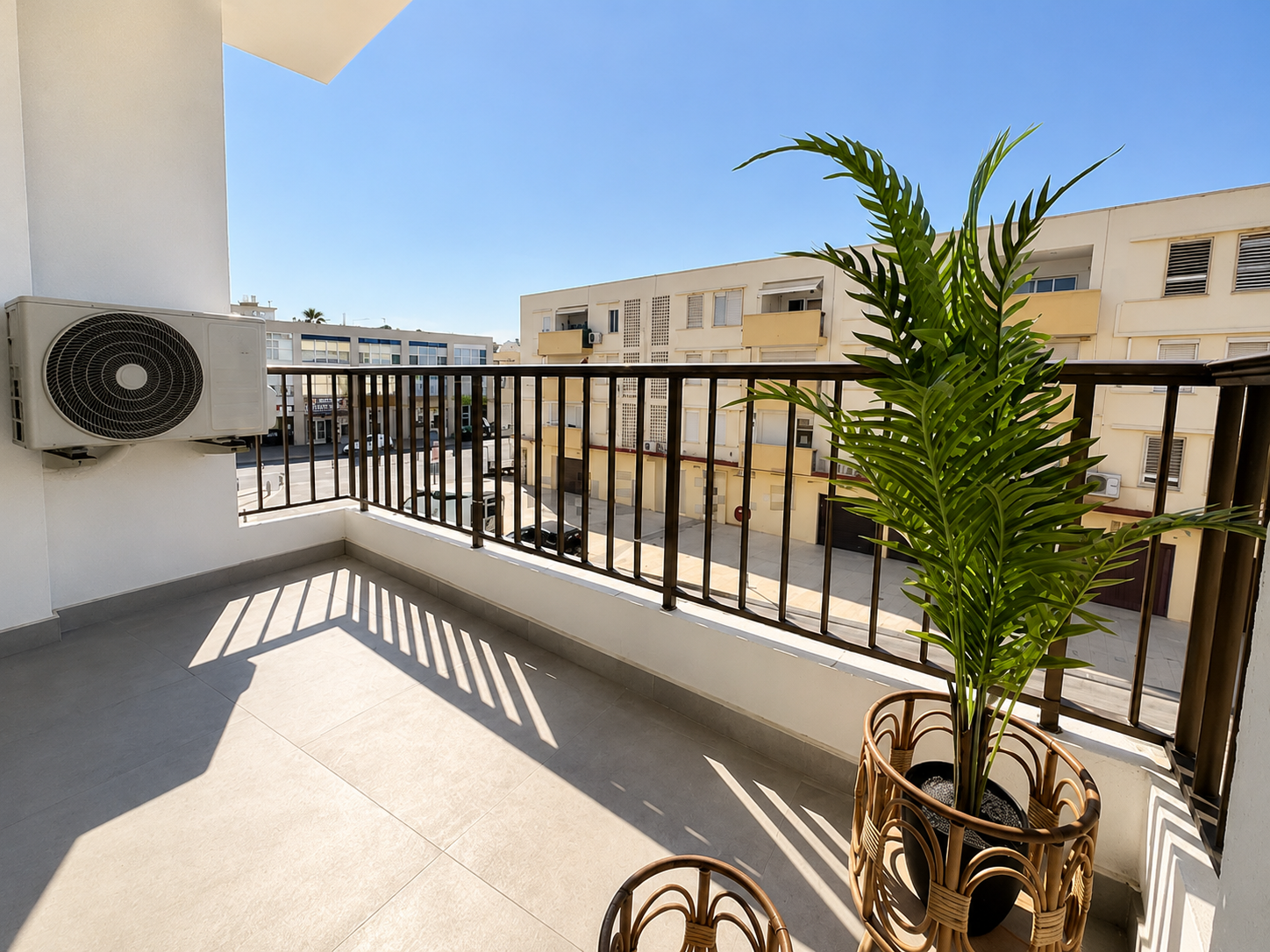 Sunny balcony with light tile flooring, a potted palm plant in a rattan stand, and a view of nearby apartment buildings. [AI-generated text]