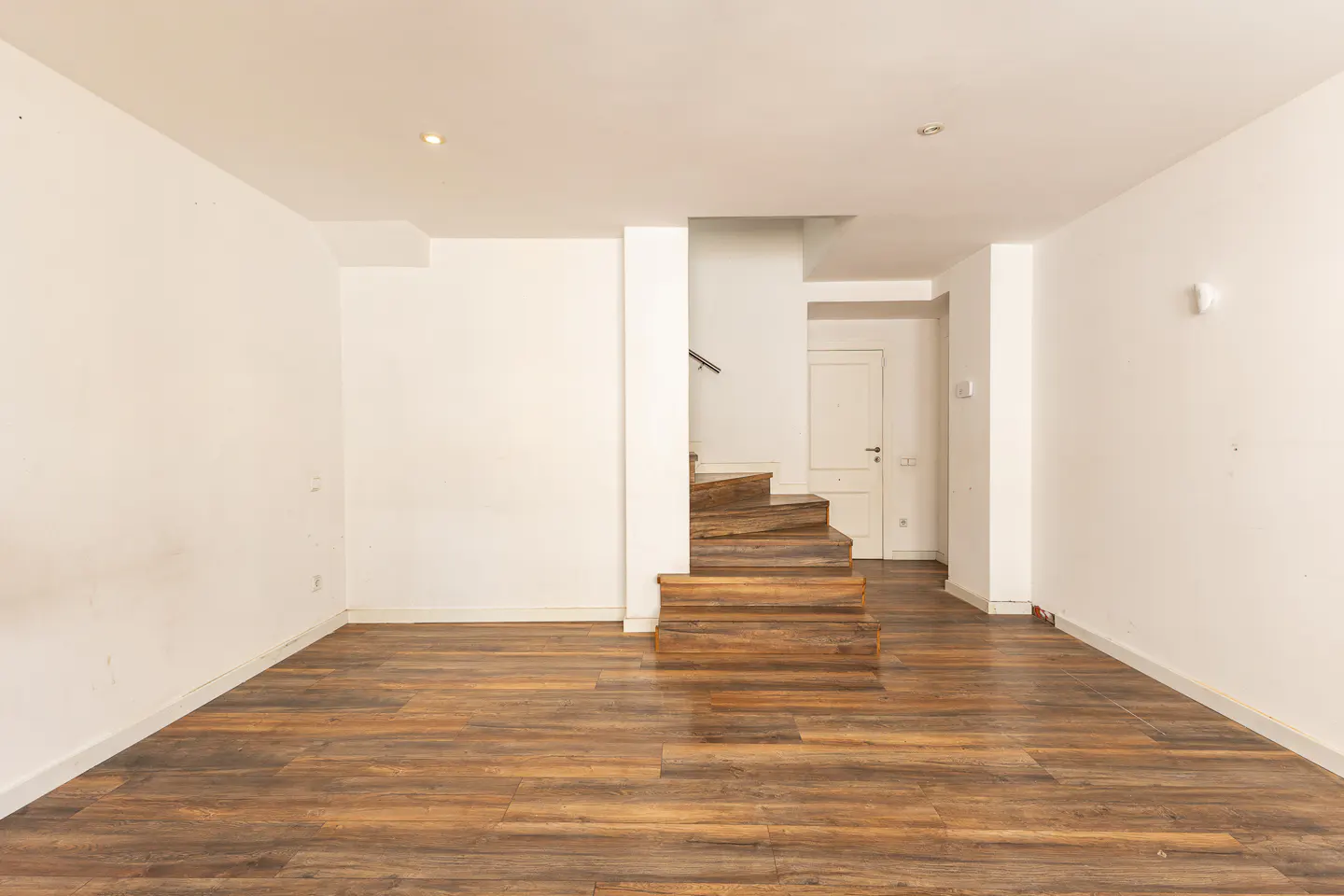 Empty room with wood-look flooring, white walls, and a wooden staircase leading to a white door.