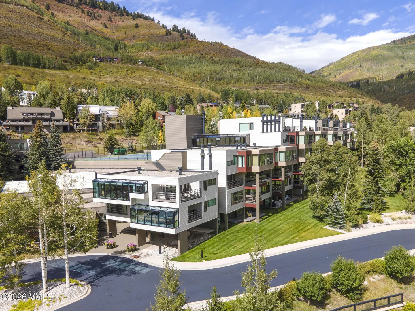 Modern white condo building with balconies, set against a mountain backdrop with green trees and blue sky.