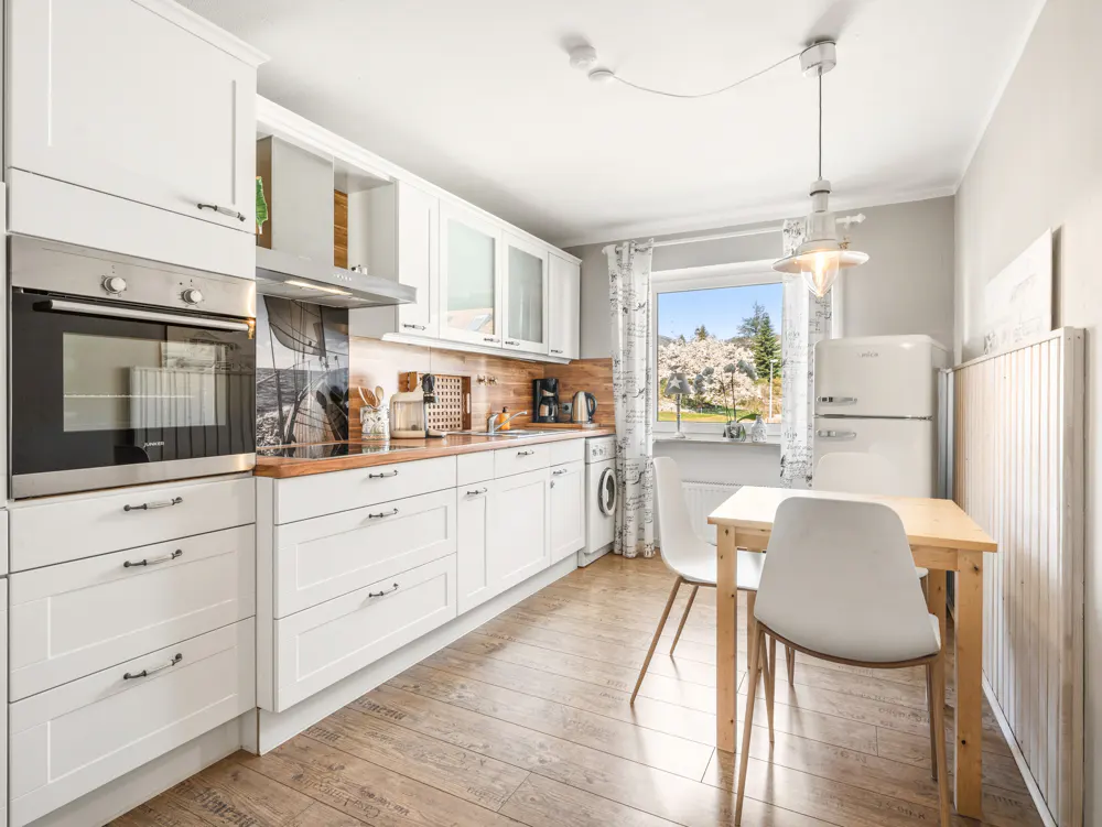 Bright kitchen with white cabinets, wood countertops, and wood floors. A table with white chairs sits near a window with a view of trees.