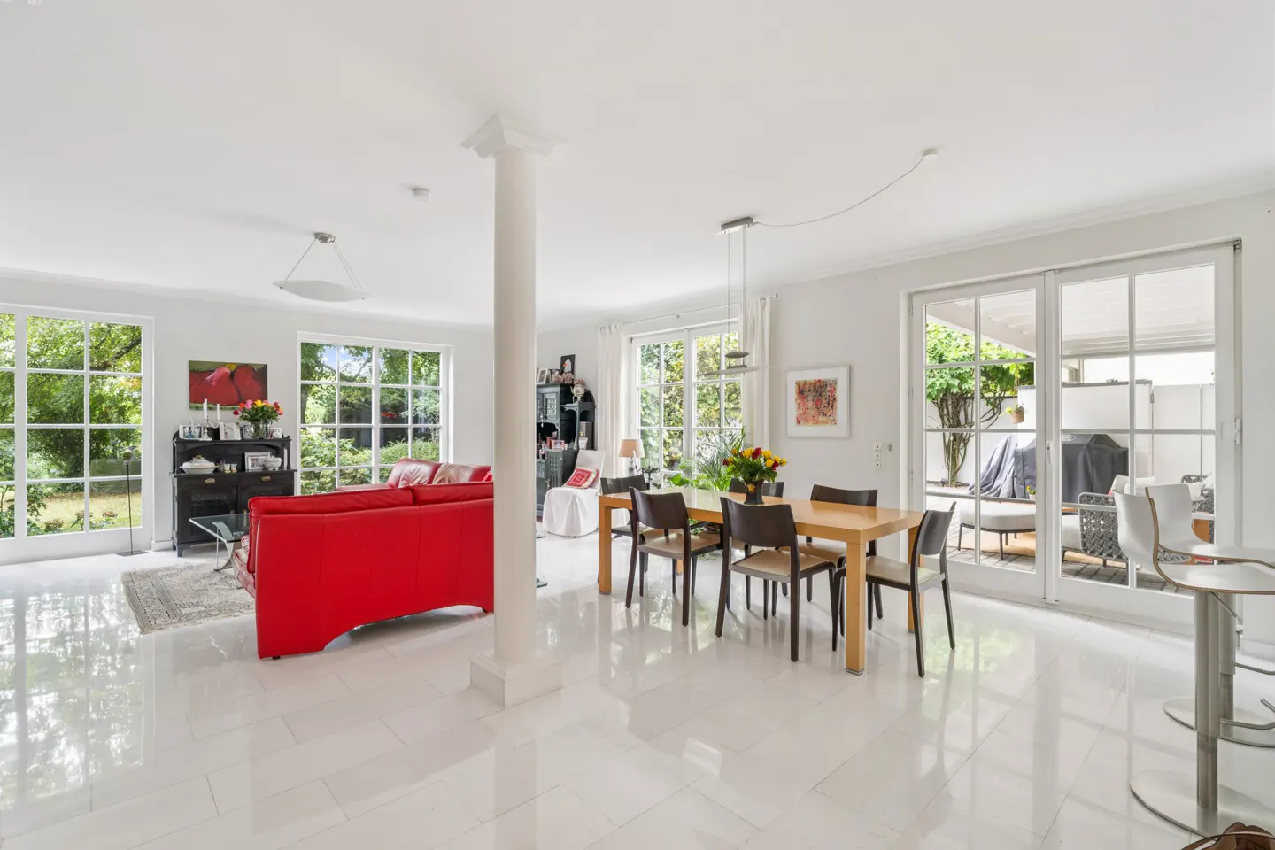 Bright, open-plan living and dining area with white walls, marble floors, and large windows overlooking a garden. A red sofa and wooden dining table are visible.