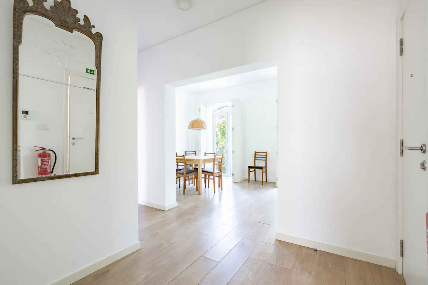 Hallway with light wood floors and white walls. A mirror hangs on the left wall, reflecting a fire extinguisher. A dining room is visible through an archway.