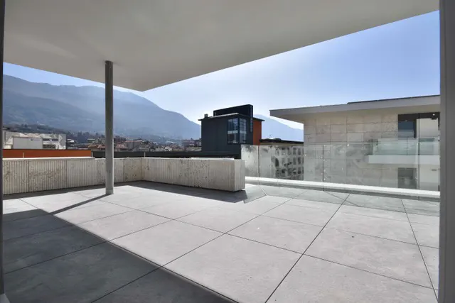 Modern rooftop terrace with gray tiles, concrete walls, and glass railings. Mountains and city buildings are visible in the background.