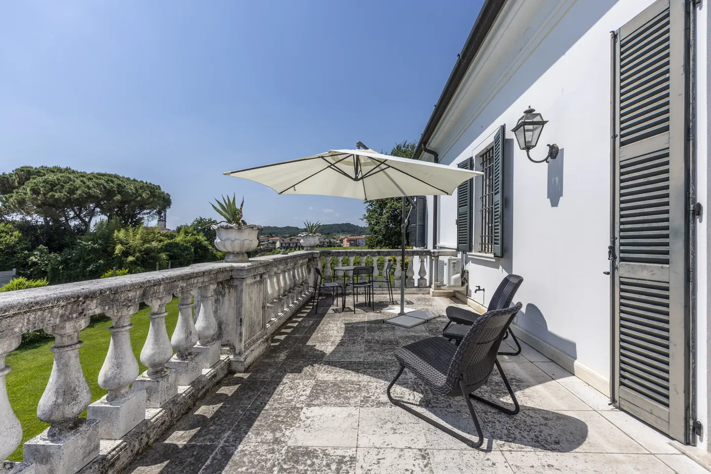 A sunny stone terrace with a white umbrella, black wicker chairs, and a classic balustrade overlooking a green garden.