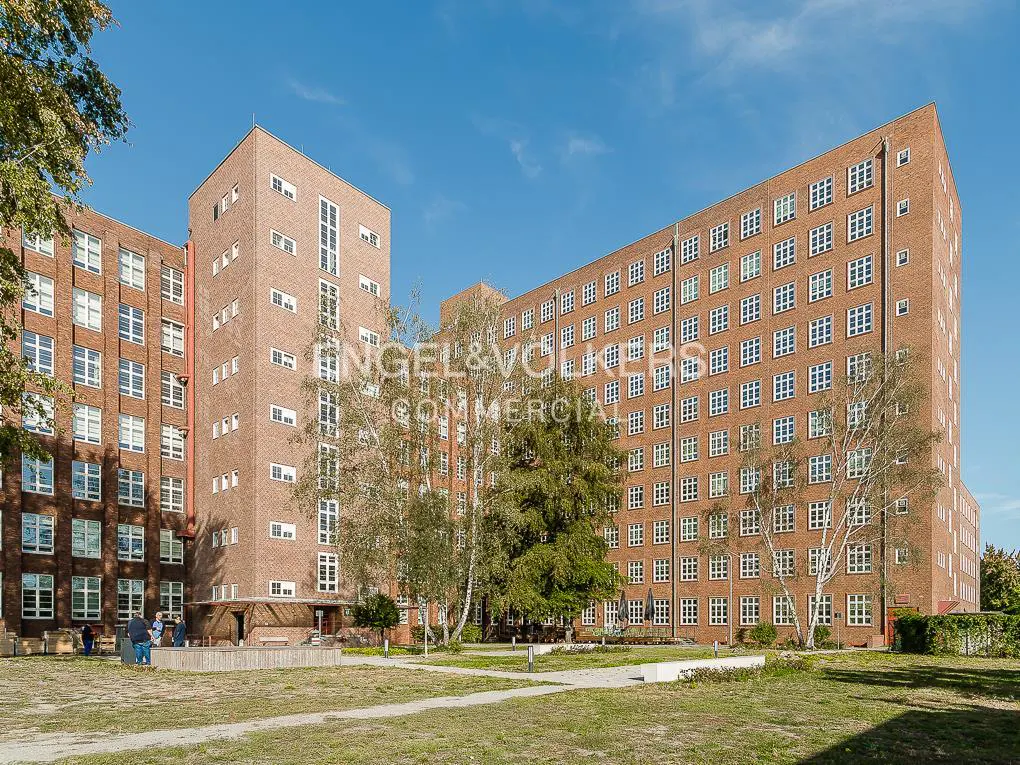 Exterior of a large, multi-story brick apartment building with many windows, trees, and a grassy lawn in front.