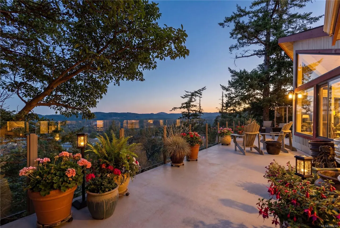 Outdoor patio with potted plants, wooden chairs, and glass railing overlooking a scenic view at dusk.
