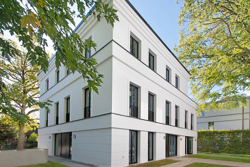 Three-story white building with black window frames, surrounded by green trees and grass.