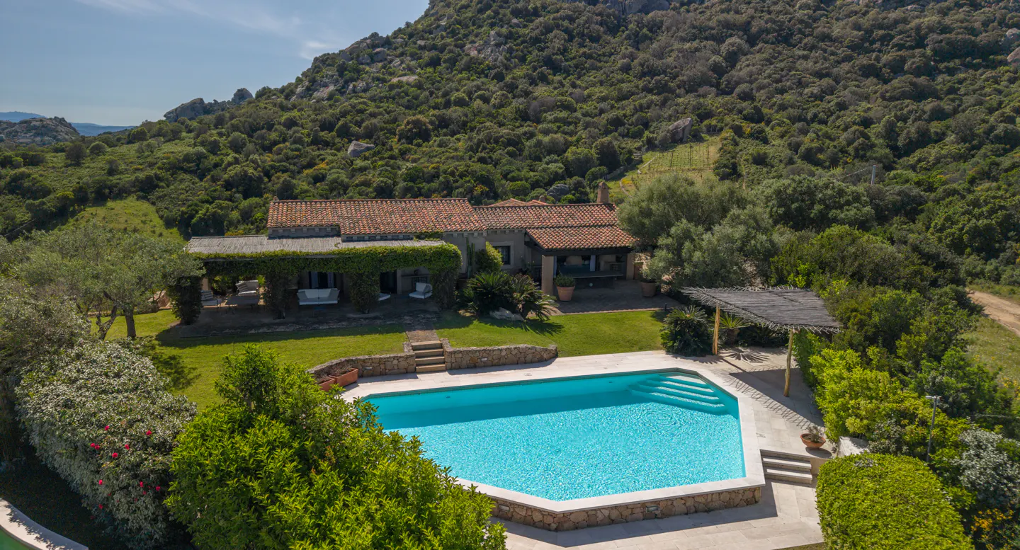 Aerial view of a luxury villa with a pool, surrounded by lush greenery and a mountain backdrop. The villa has a red tile roof.