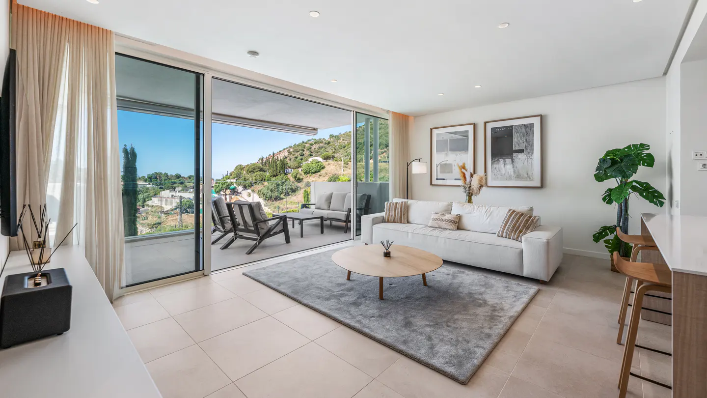 Bright living room with a white sofa, round wood table on a gray rug, and a balcony with a view of green hills.