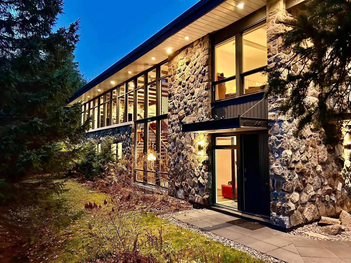 Exterior view of a stone house with large windows and a spiral staircase visible inside. The front door is open, revealing a glimpse of the interior.