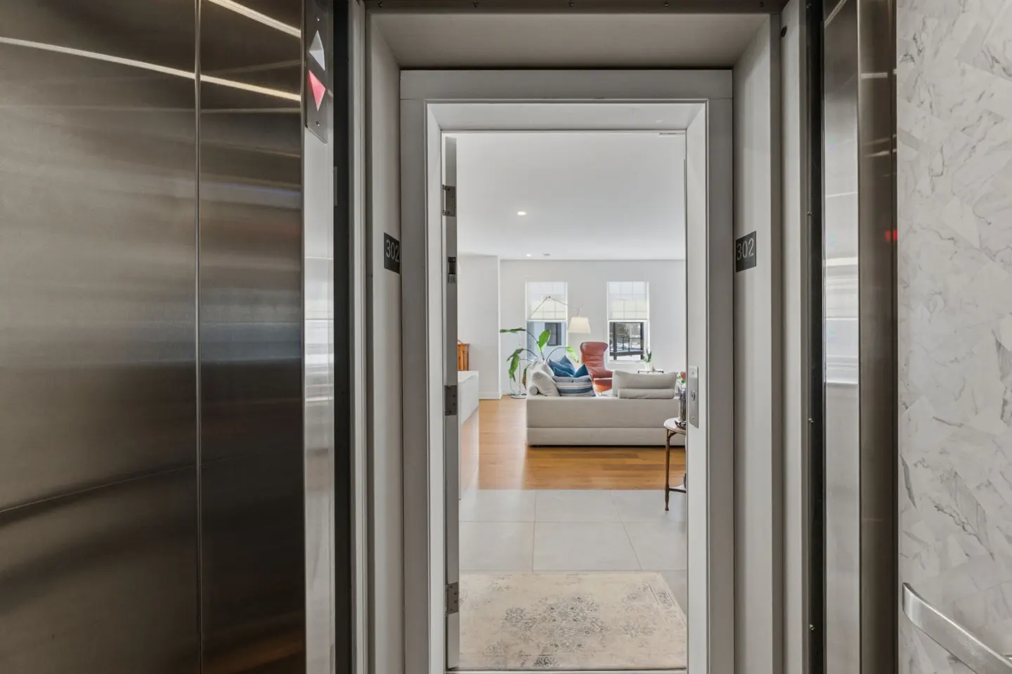 View from elevator of a living room. Stainless steel doors open to a white room with a couch, wood floors, and windows.