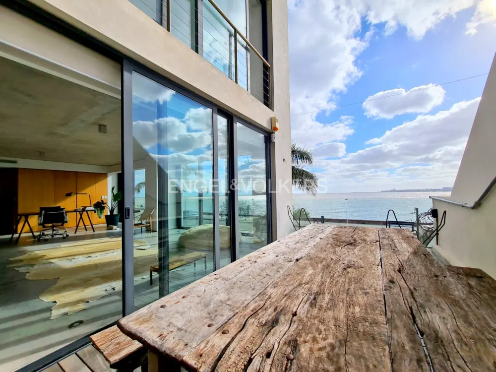 Exterior view of a modern home with a weathered wood table on the patio overlooking the ocean. Sliding glass doors reflect the blue sky.