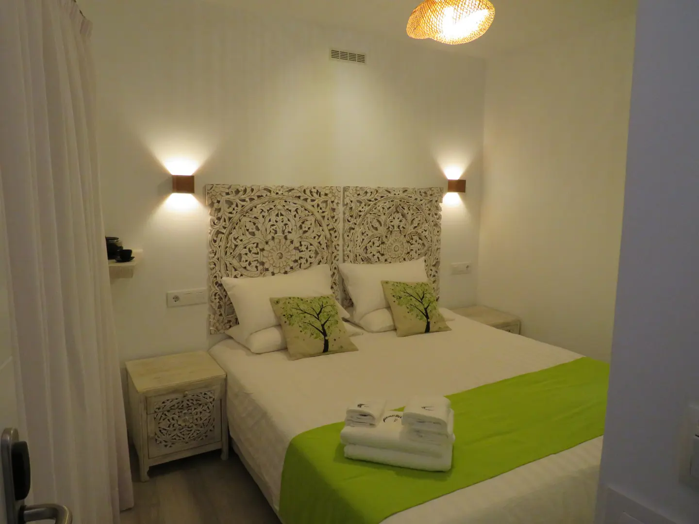Bright bedroom with a carved white headboard, white linens, and a green runner. Tree-patterned pillows and carved nightstands add detail.