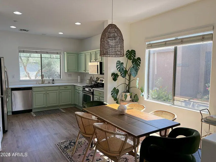 A bright, open kitchen and dining area with green cabinets, a wooden table, and wicker chairs. A large monstera plant sits near a window.