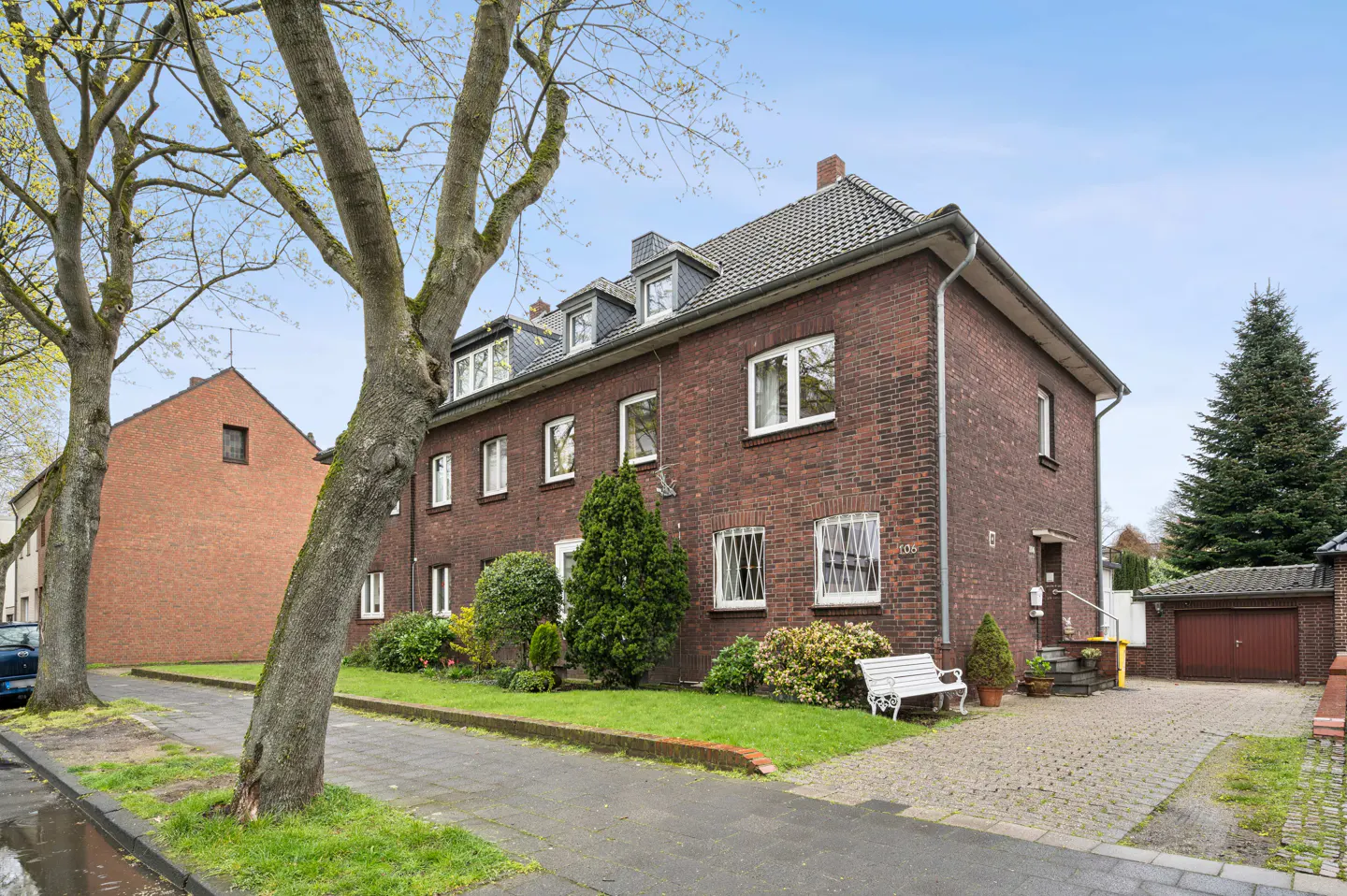 A three-story red brick apartment building with white windows and a gray roof. A white bench sits on the lawn.