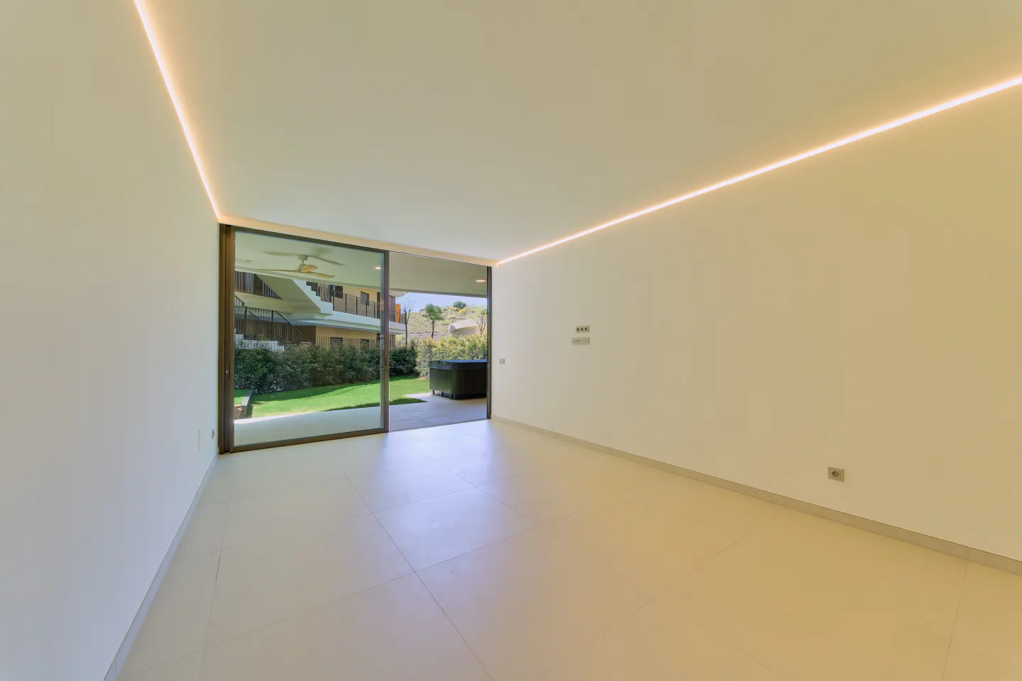 Bright, empty room with white walls, tile floor, and recessed lighting. Sliding glass doors open to a patio, lawn, and house.