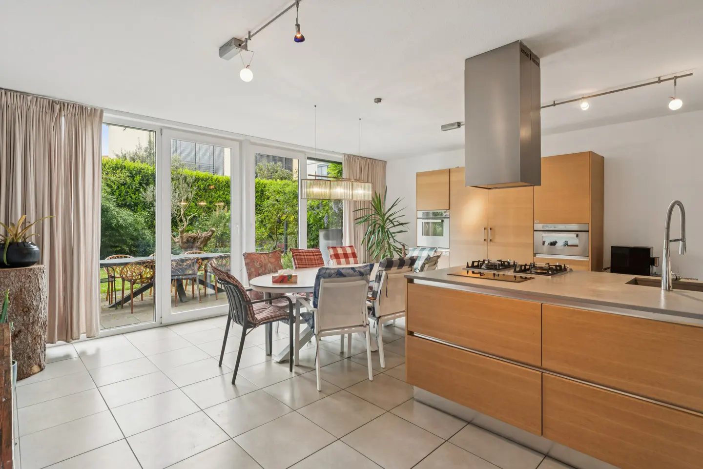 Modern kitchen and dining area with light wood cabinets, a central island, and floor-to-ceiling windows overlooking a garden.