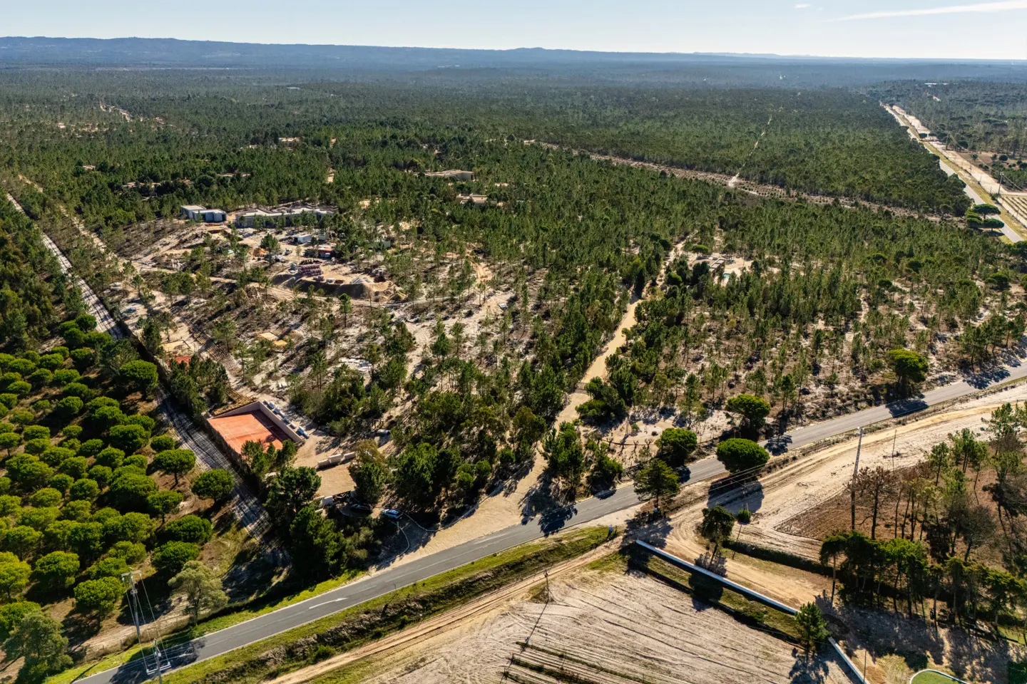Aerial view of a property with a tennis court, buildings, and a dense forest in the background. A road runs through the landscape.