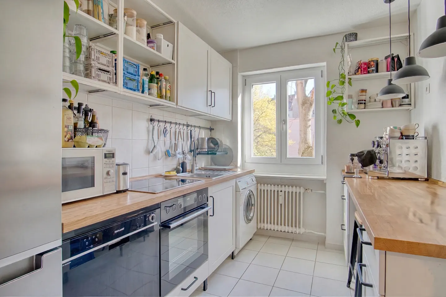 Bright kitchen with white cabinets, wood countertops, and white tiled floors. A window overlooks a tree. Appliances include a black oven and a microwave.