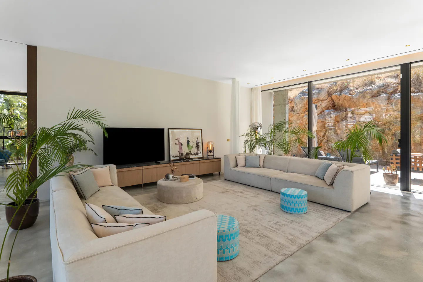Bright living room with beige sofas, a round ottoman, and a large TV. Sliding glass doors reveal a rocky landscape. Plants add a touch of nature.