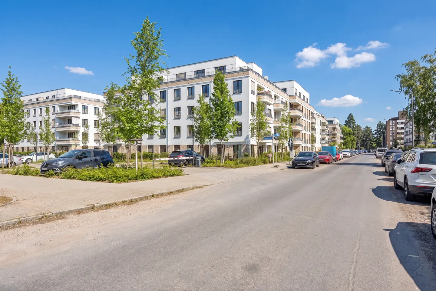 Street view of modern white apartment buildings with balconies, cars parked along the road, and green trees under a blue sky.