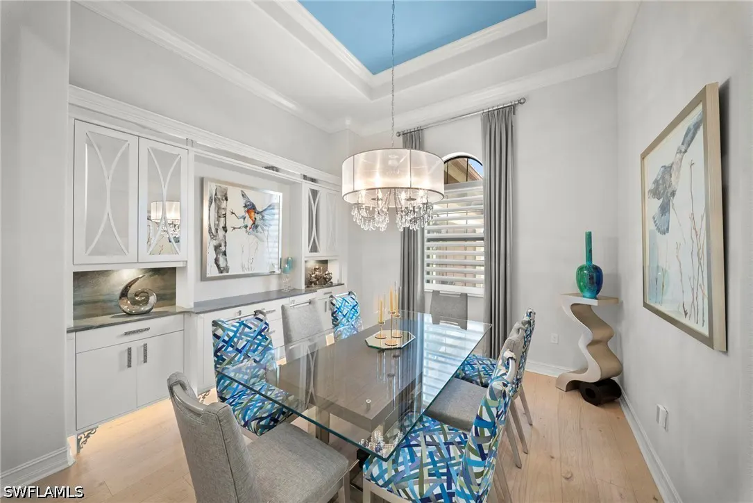 A dining room with a glass table, blue patterned chairs, and a white cabinet with artwork. A chandelier hangs from a blue ceiling accent.