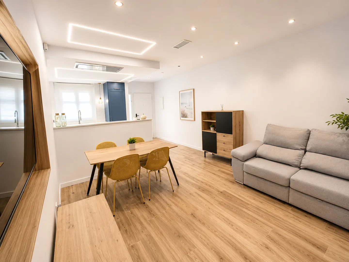 Bright, modern apartment interior with wood floors, a gray sofa, a dining table with yellow chairs, and a partial view of the kitchen.