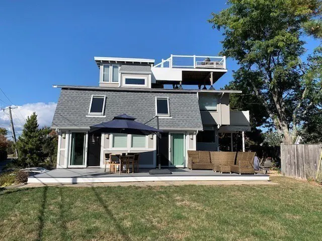 Exterior view of a two-story house with a gray roof, a deck with outdoor furniture, and a blue umbrella.