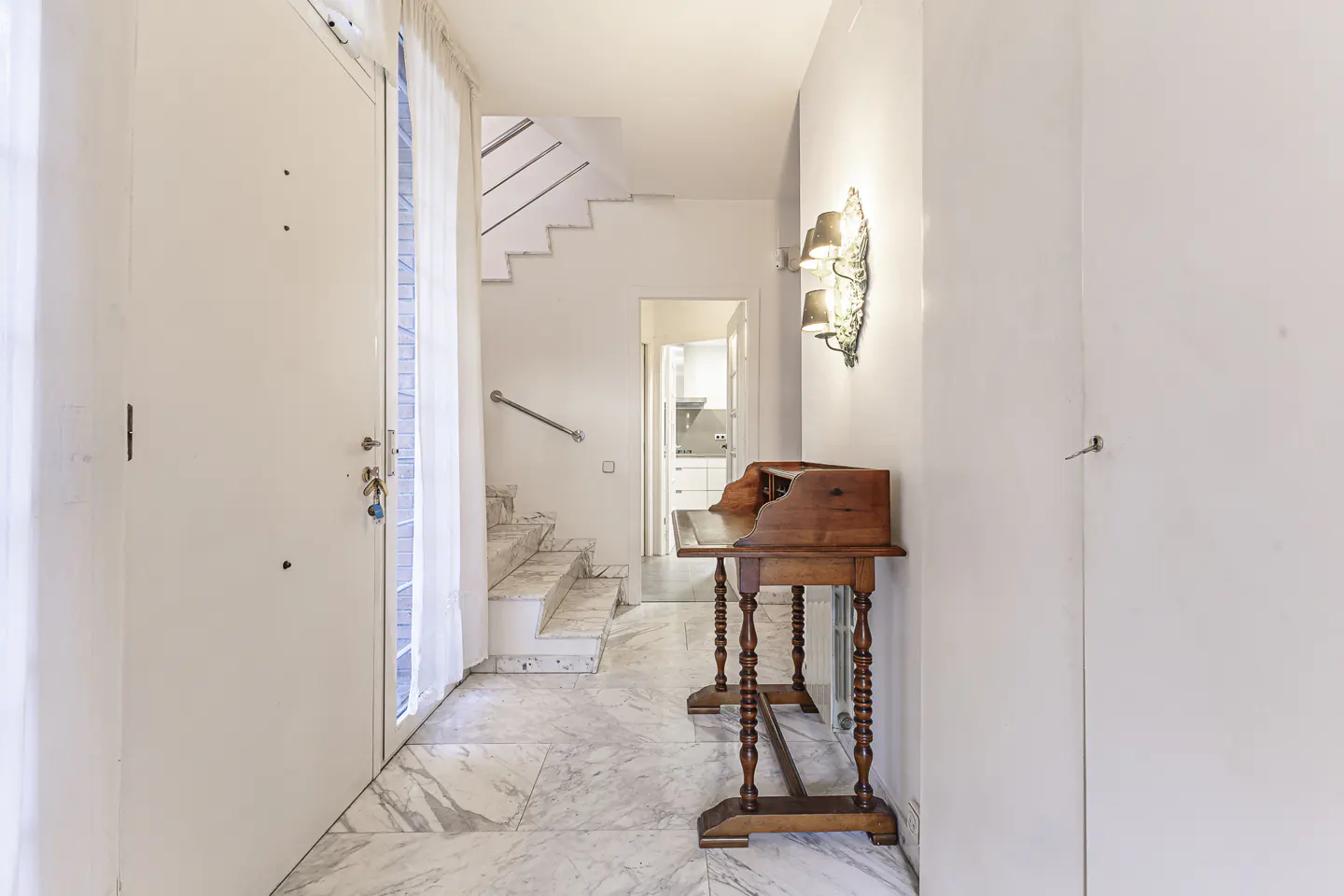 Entryway with marble floors, white walls, and a wooden desk. Stairs lead up, and a doorway shows a glimpse of the kitchen.