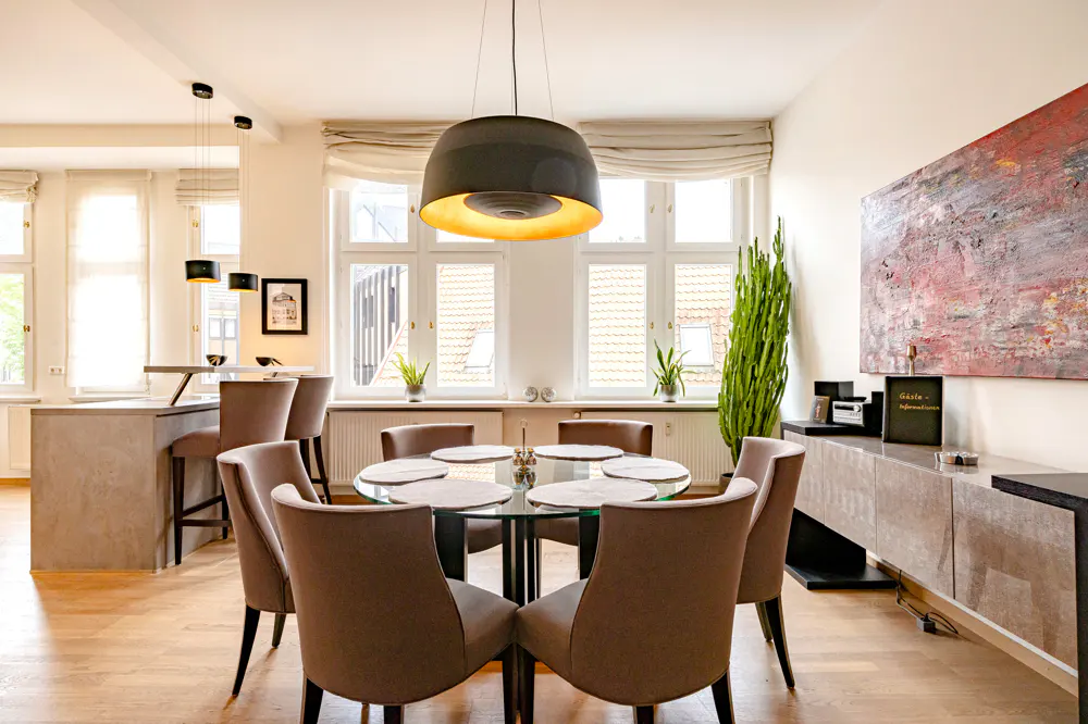Bright dining room with a glass table, brown chairs, and a large black pendant light. Windows with beige blinds and a modern painting are visible.