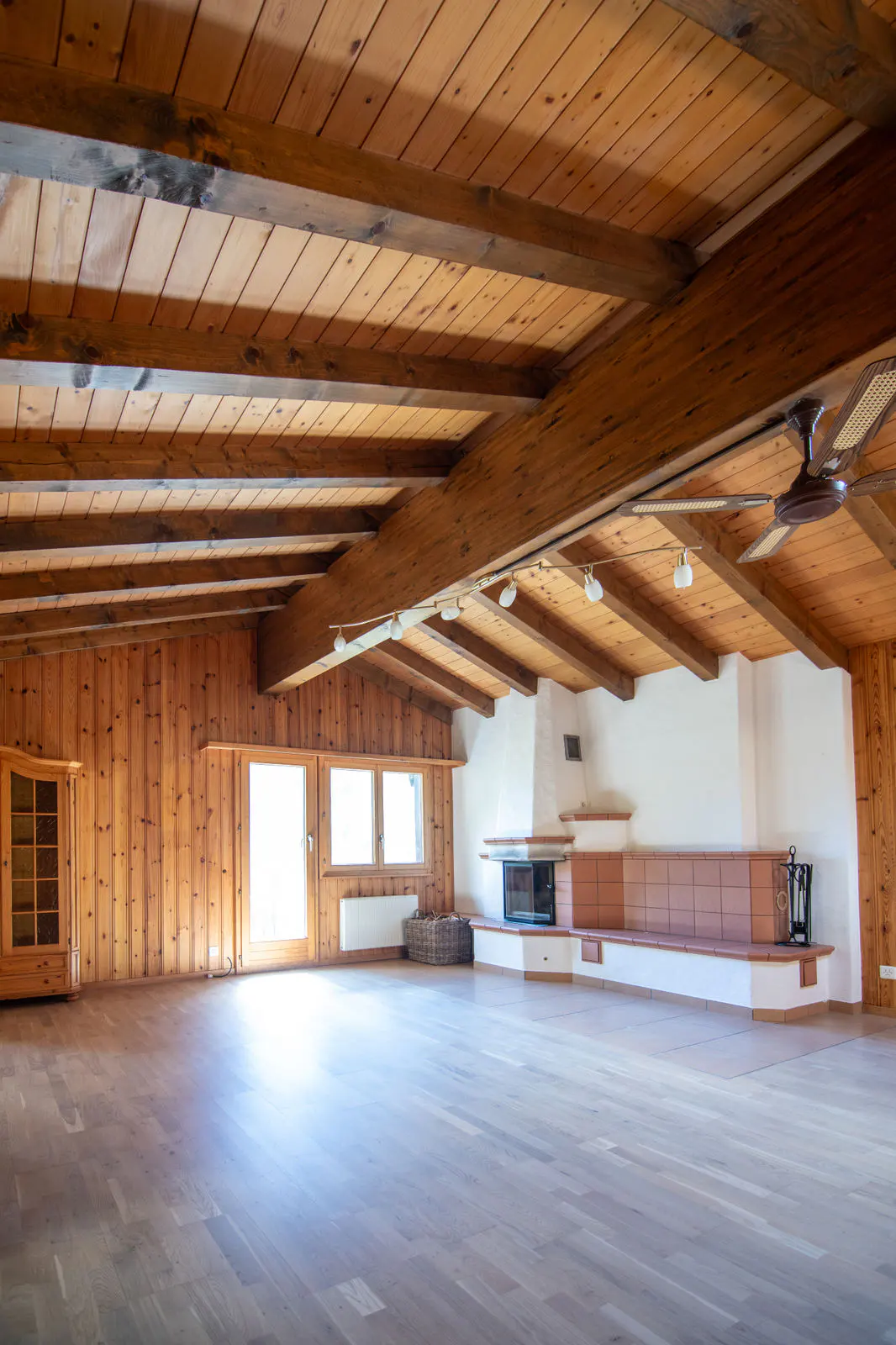 Spacious room with wood-paneled walls, exposed beam ceiling, and light wood floors. A fireplace and windows provide natural light.