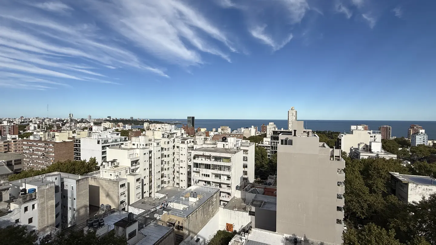 Cityscape view with white buildings, green trees, and blue sky with wispy clouds. Ocean visible in the background.