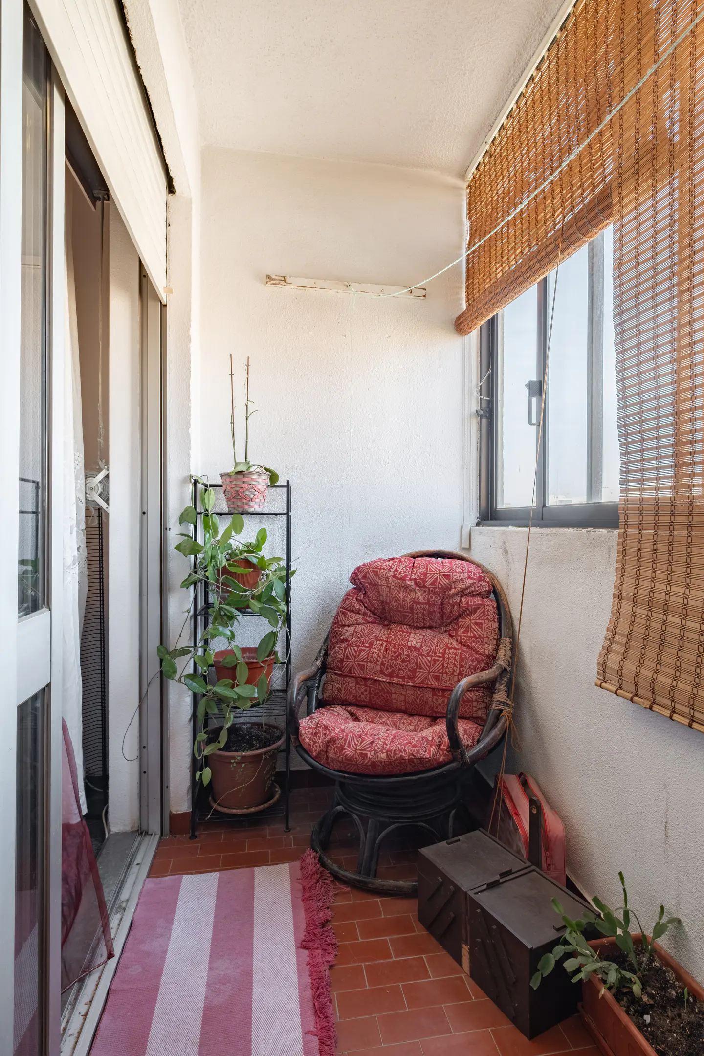 A cozy balcony with a red-cushioned chair, potted plants on a rack, and a striped rug on red tile flooring. A bamboo blind covers the window.