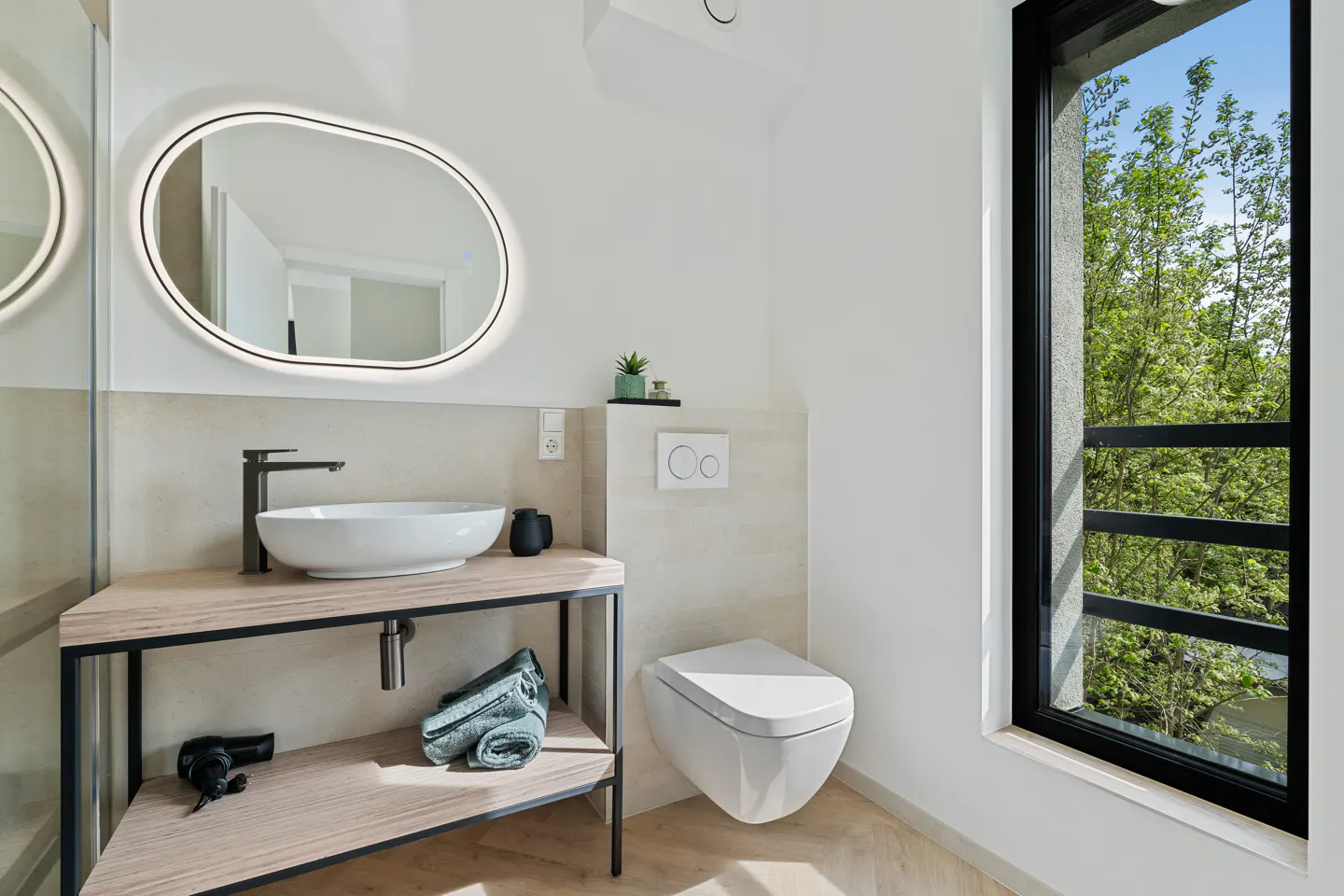 Bright bathroom with a white sink on a wood and metal stand, a toilet, and a window with green trees outside.