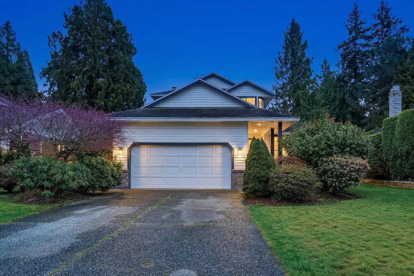 Exterior shot of a two-story white house with a two-car garage, green lawn, and trees at dusk.