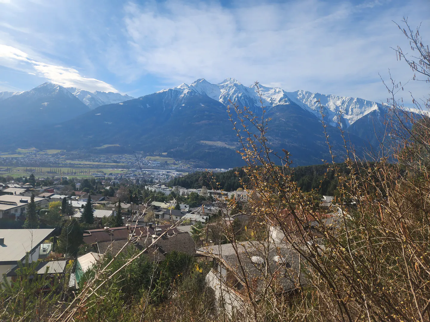 View of a town nestled below snow-capped mountains under a blue sky with scattered clouds. Foreground shows bare branches.