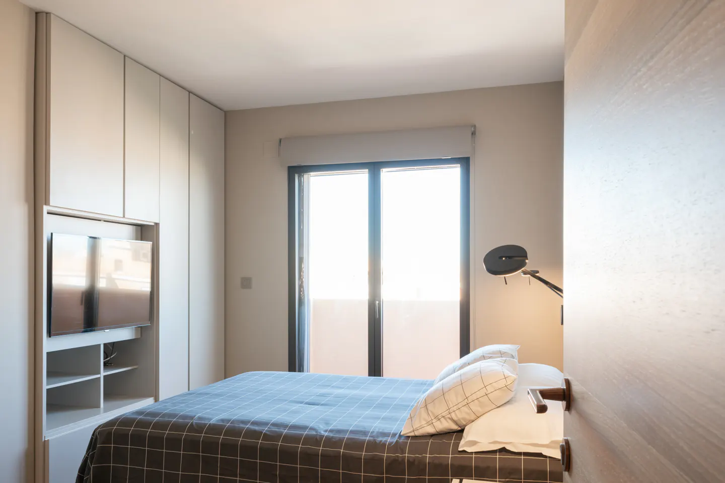 A modern bedroom with a gray plaid bedspread, white pillows, and a black adjustable lamp. A TV is mounted in a gray cabinet. Bright light shines through the window.