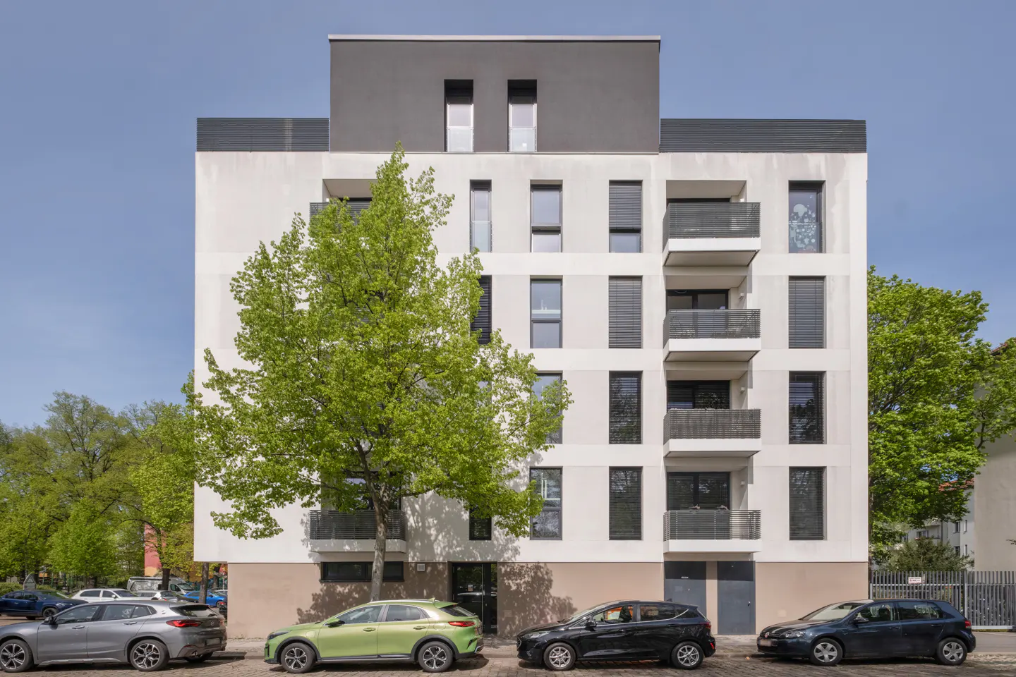Modern apartment building with white facade, gray top floor, and balconies. Cars are parked in front, and a green tree is on the left.