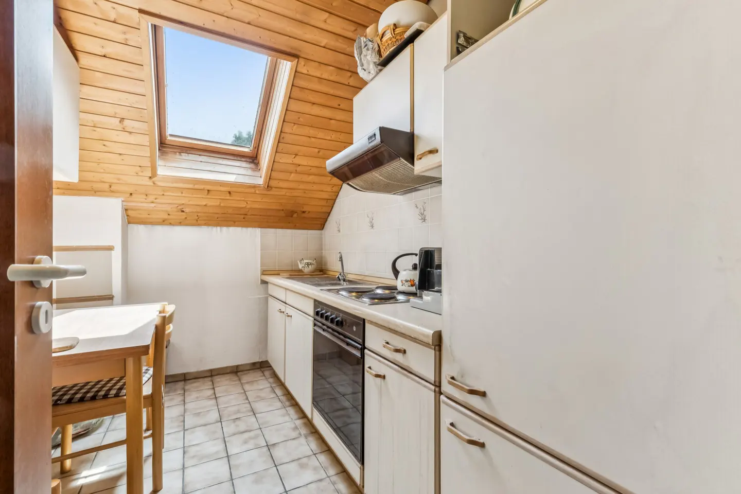 Attic kitchen with white cabinets, tiled floor, wooden sloped ceiling, skylight, and a small dining table with chairs.
