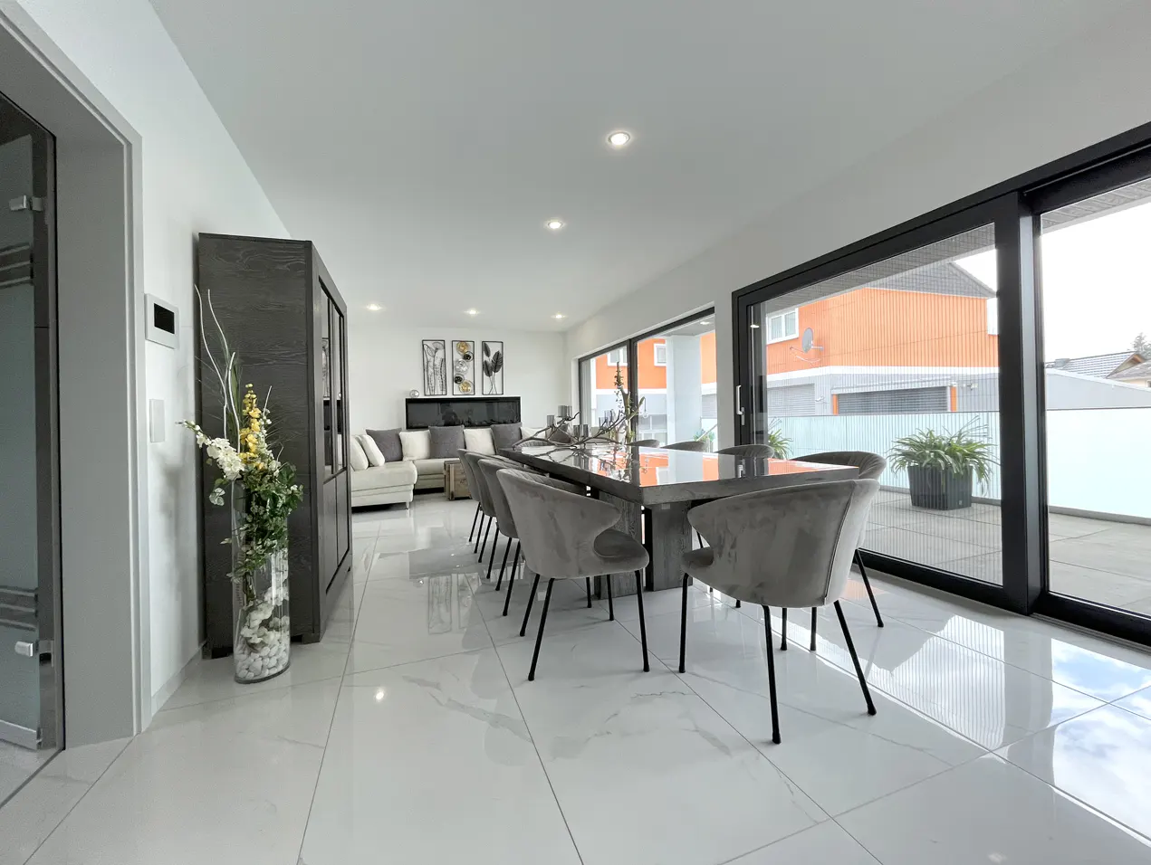 A modern, open-concept dining and living room with white marble floors. A long gray dining table with velvet chairs sits near sliding glass doors.