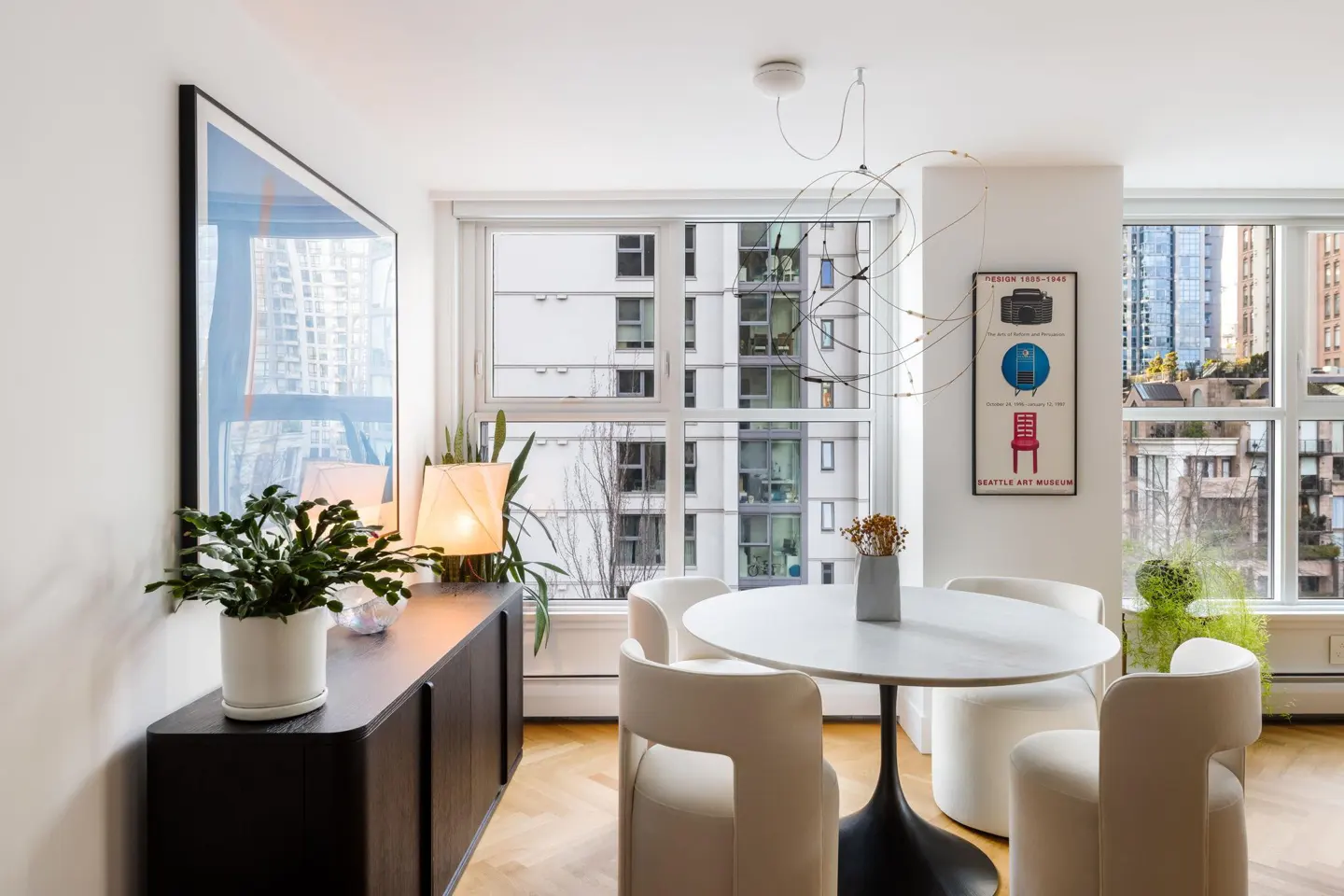 Bright dining area with a round white table, four white chairs, and a black sideboard. Large windows offer city views. Modern art and plants add color.