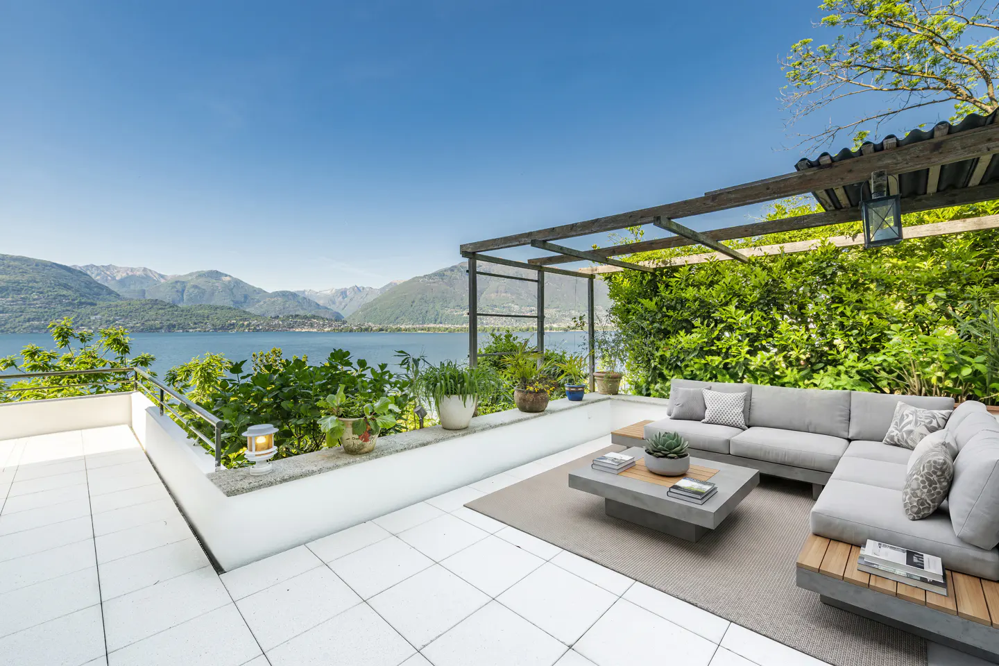 Outdoor patio with gray sectional sofa, concrete table, and lake view. White tile floor, green plants, and blue sky.