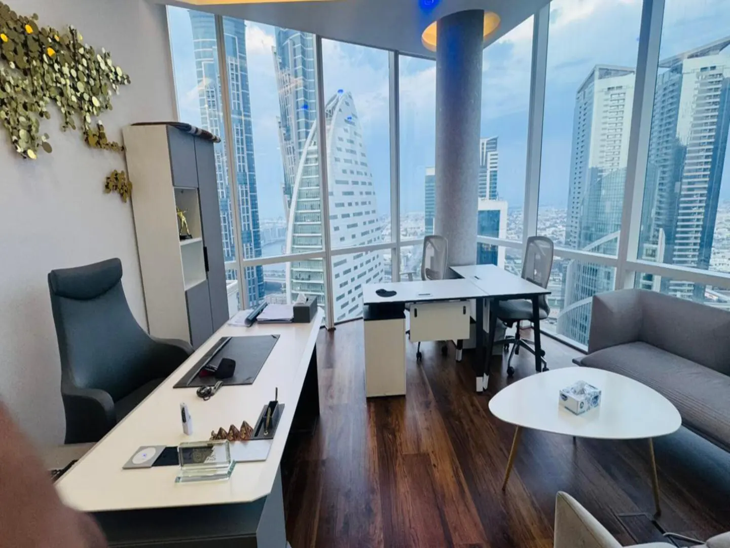 A modern office with floor-to-ceiling windows overlooking a cityscape. Desks, chairs, and a sofa furnish the space.