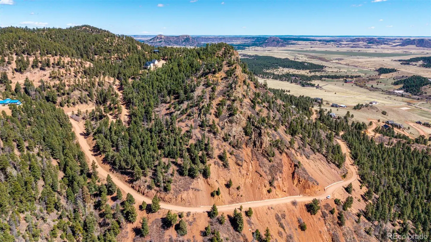 Aerial view of a red rock mountain with green trees and a winding dirt road under a blue sky.