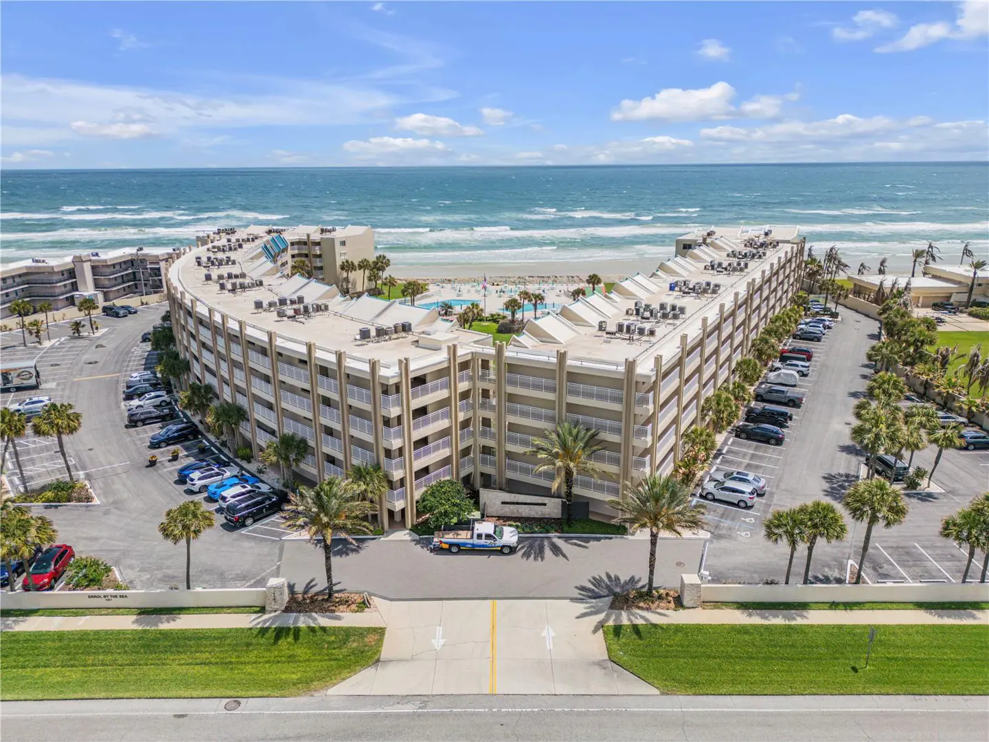 Aerial view of a curved, multi-story condo building with a pool, parking lot, and ocean backdrop under a blue sky.