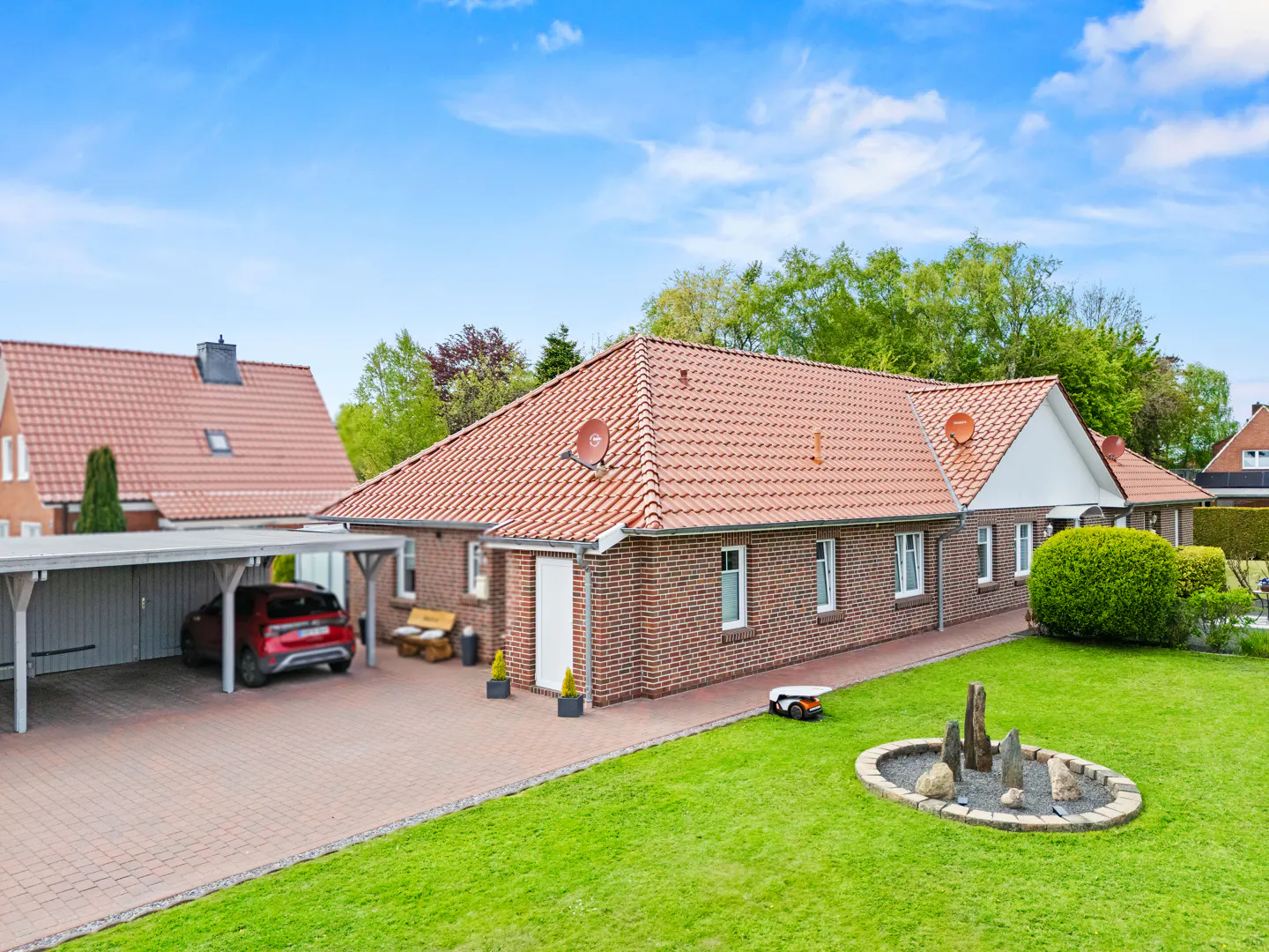 Brick house with a red tile roof, a carport with a red car, and a green lawn with a robotic lawnmower.
