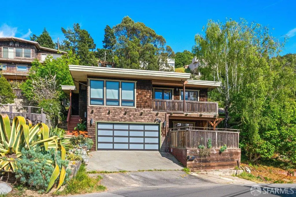 Two-story brown shingle house with a blue-paneled garage and wooden balconies on a sunny day.